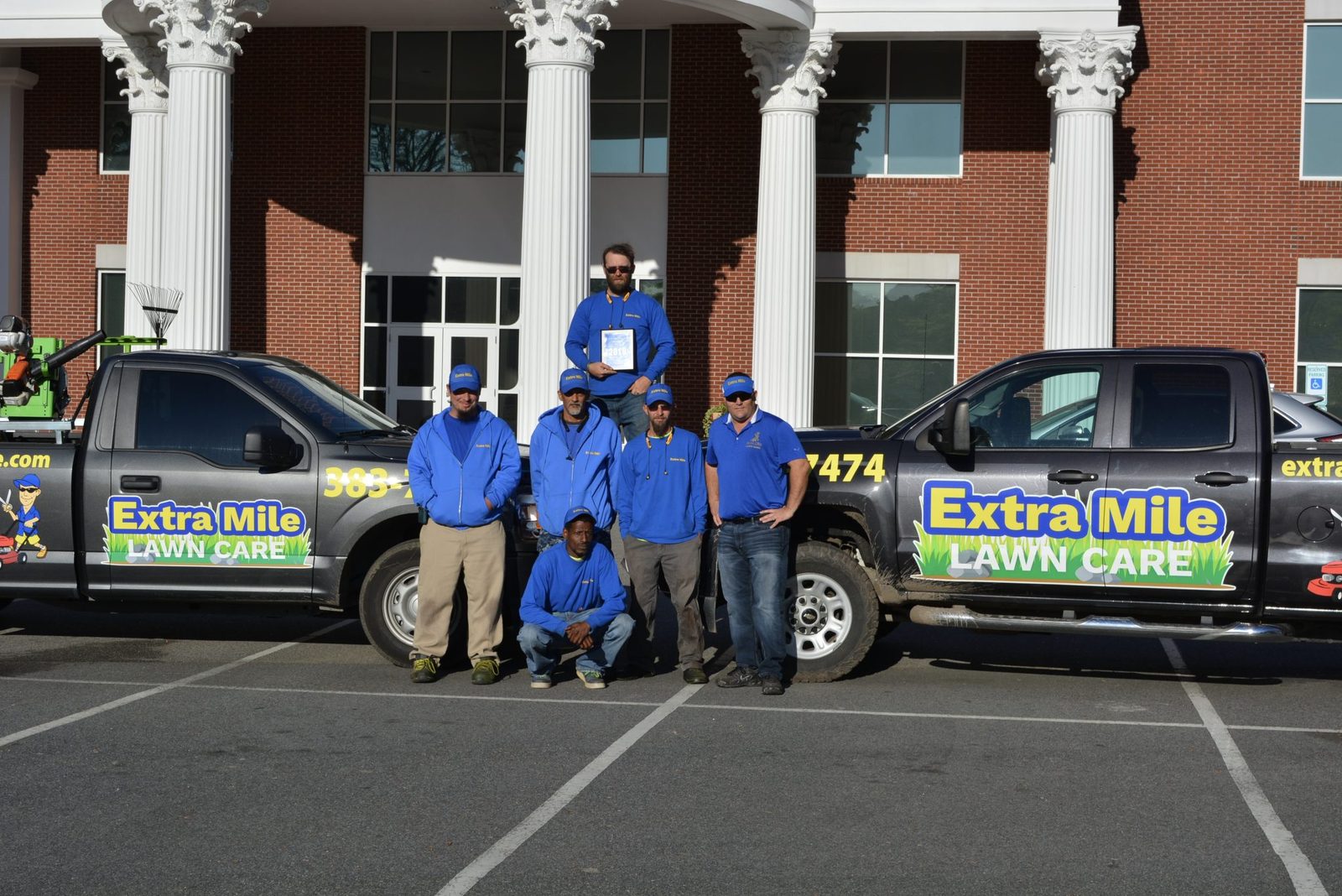 The Extra Mile Lawn Care crew in branded blue uniforms standing between two company trucks loaded with mowers and blowers.